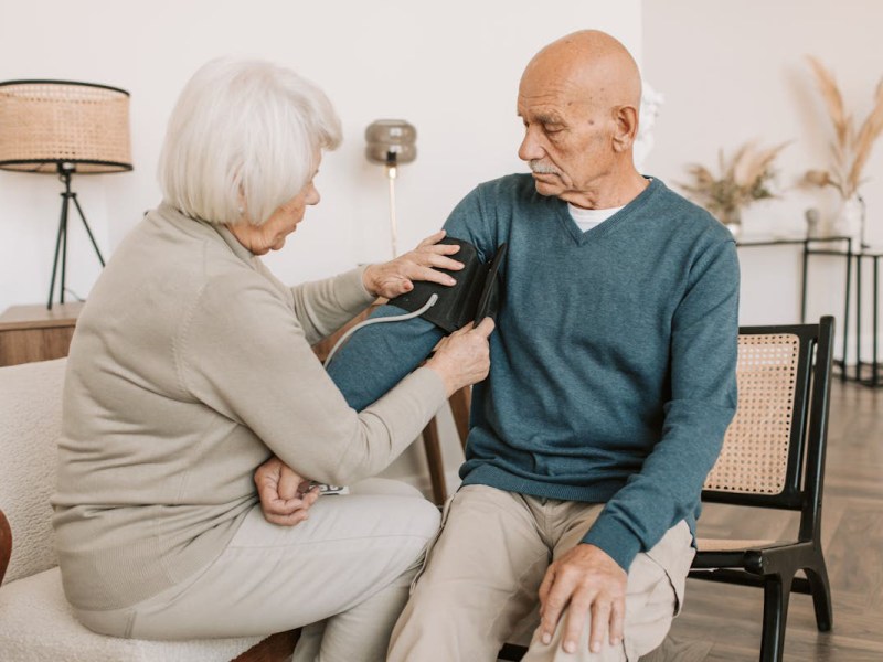 Älteres Ehepaar sitzt auf einem Sofa. Sie legt ihm eine Manschette zum Blutdruckmessen an.