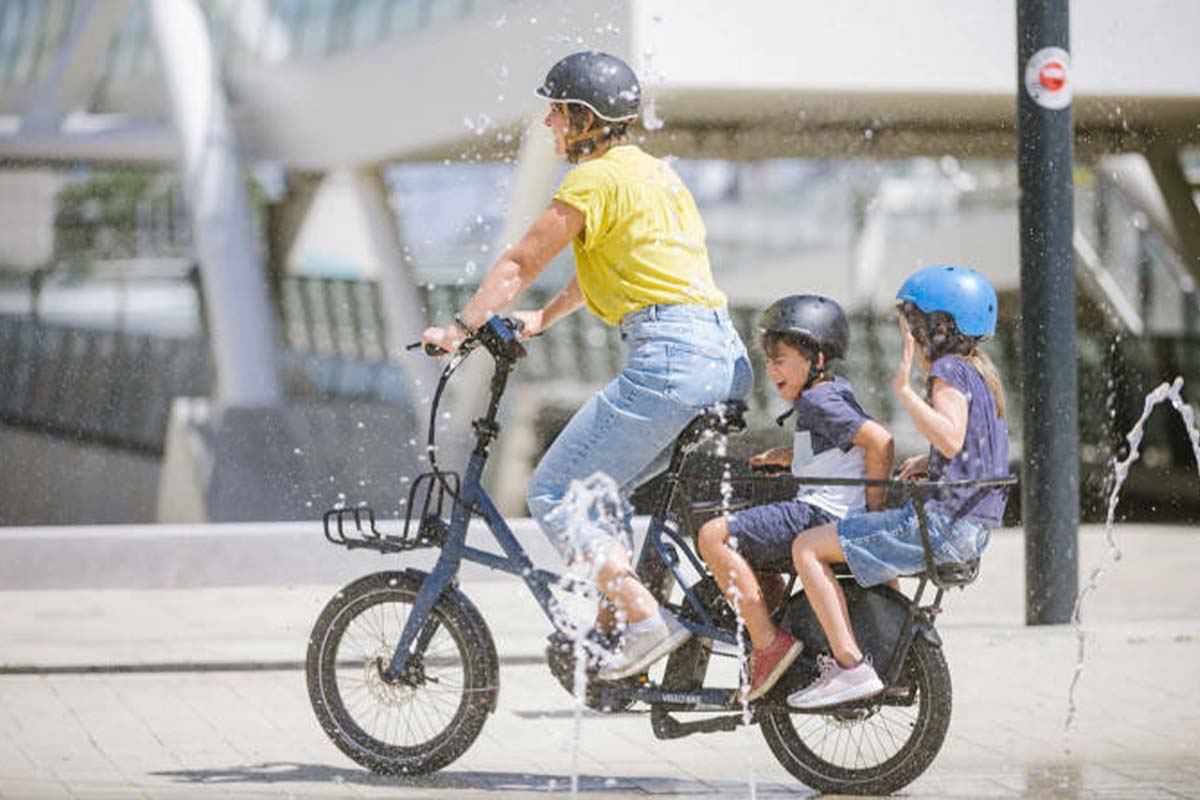 Kompaktes E-Lastenrad Sub von Vello, Frau fährt damit durch Stadt, zwei Kinder sind auf dem Gepäckträger