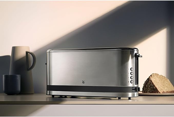 Der silberne Langschlitz-Toaster von WMF steht mittig im Bild auf einer Anrichte, links daneben befinden sich Krug und Becher, rechts daneben ein angeschnittenes Körnerbrot auf einem Teller