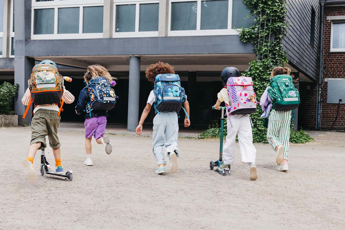 Fünf Kinder rennen in ein Schulgebäude, man sieht sie von hinten, alle tragen Ranzen