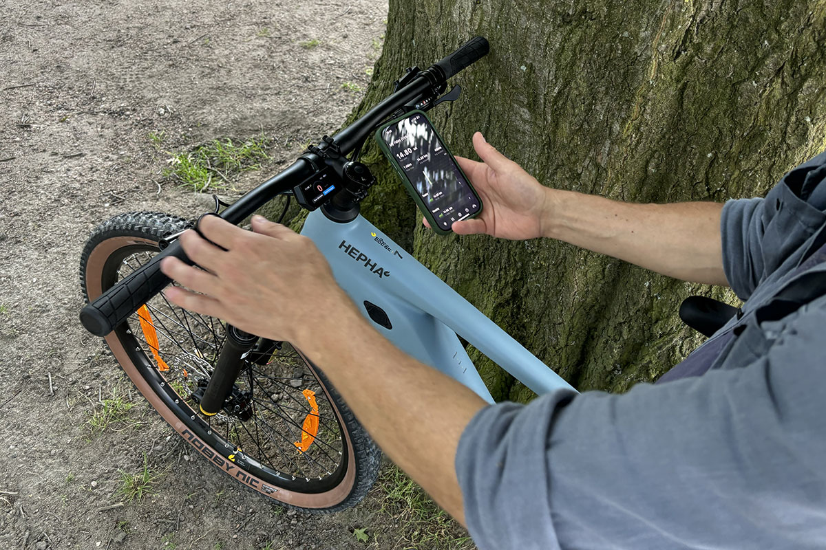 Mann verbindet Smartphone-App mit einem E-Bike. Dafür hälöt er ein Smartphone über den Lenker, des an einem Baum stehenden E-Bikes und bedient mit der anderen Hand ein Steuerungsmodul am E-Bike-Lenker.