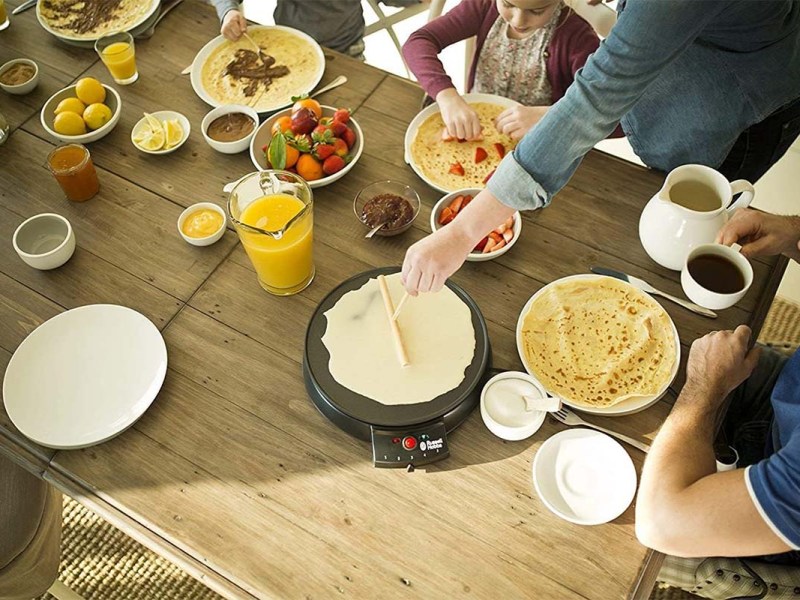 Blick auf einen Tisch, auf dem ein Crepes Maker und viele Zutaten drumherum stehen. Eine Hand verteilt mit einem Rechen Teig auf dem Gerät