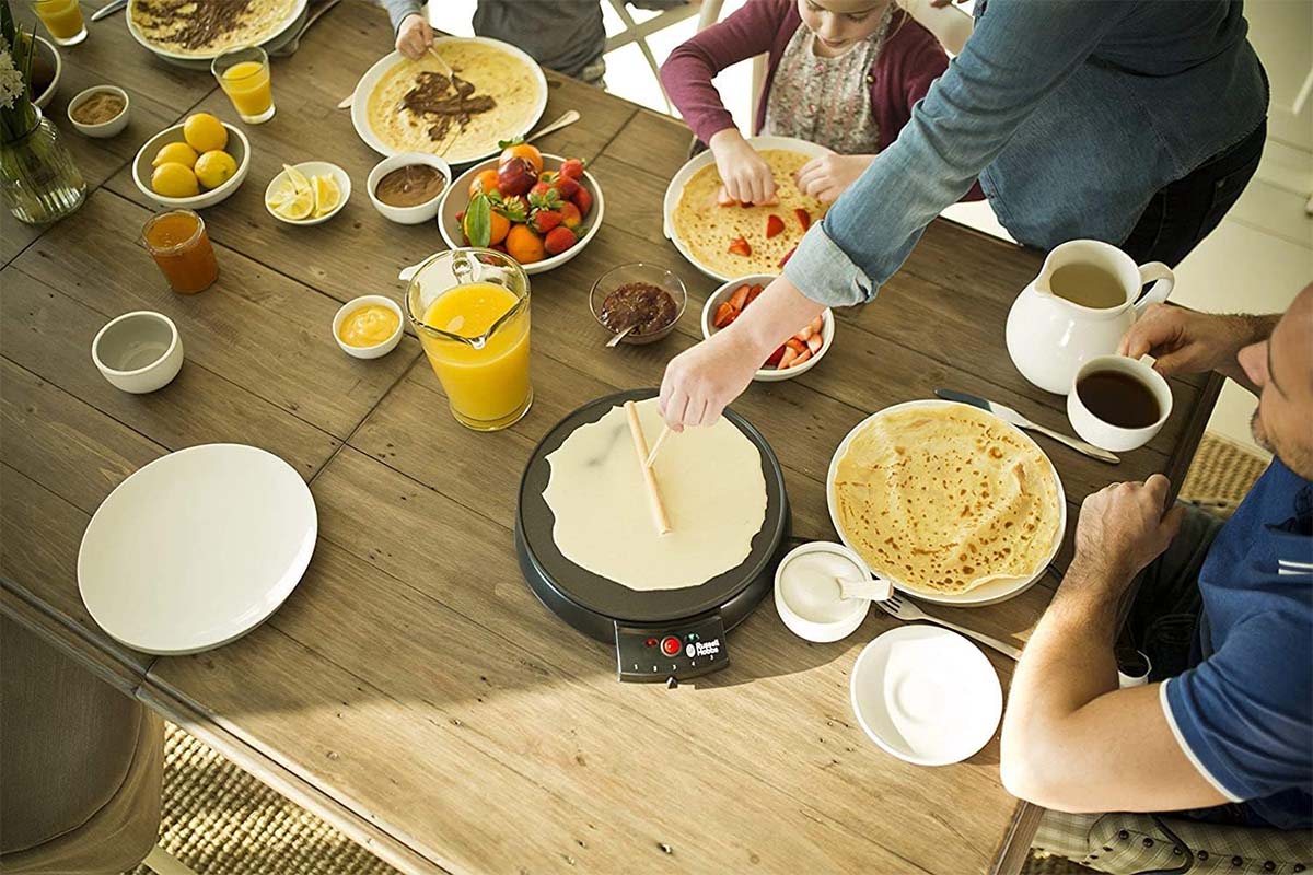 Blick auf einen Tisch, auf dem ein Crepes Maker und viele Zutaten drumherum stehen. Eine Hand verteilt mit einem Rechen Teig auf dem Gerät