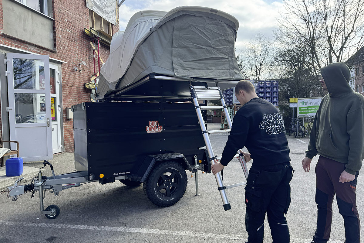 Mann zieht Leiter eines Dachzelts aus, das auf einem Amhänger montiert und aufgeklappt ist.