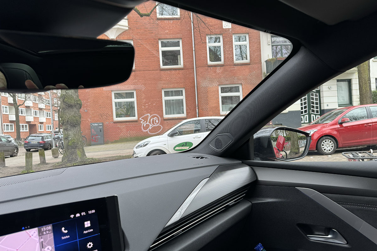 Blick schräg nach rechts durch die Frontscheibe des E-Autos Opel Astra Electric auf ein Wohnhaus in einer städtischen Umgebung.