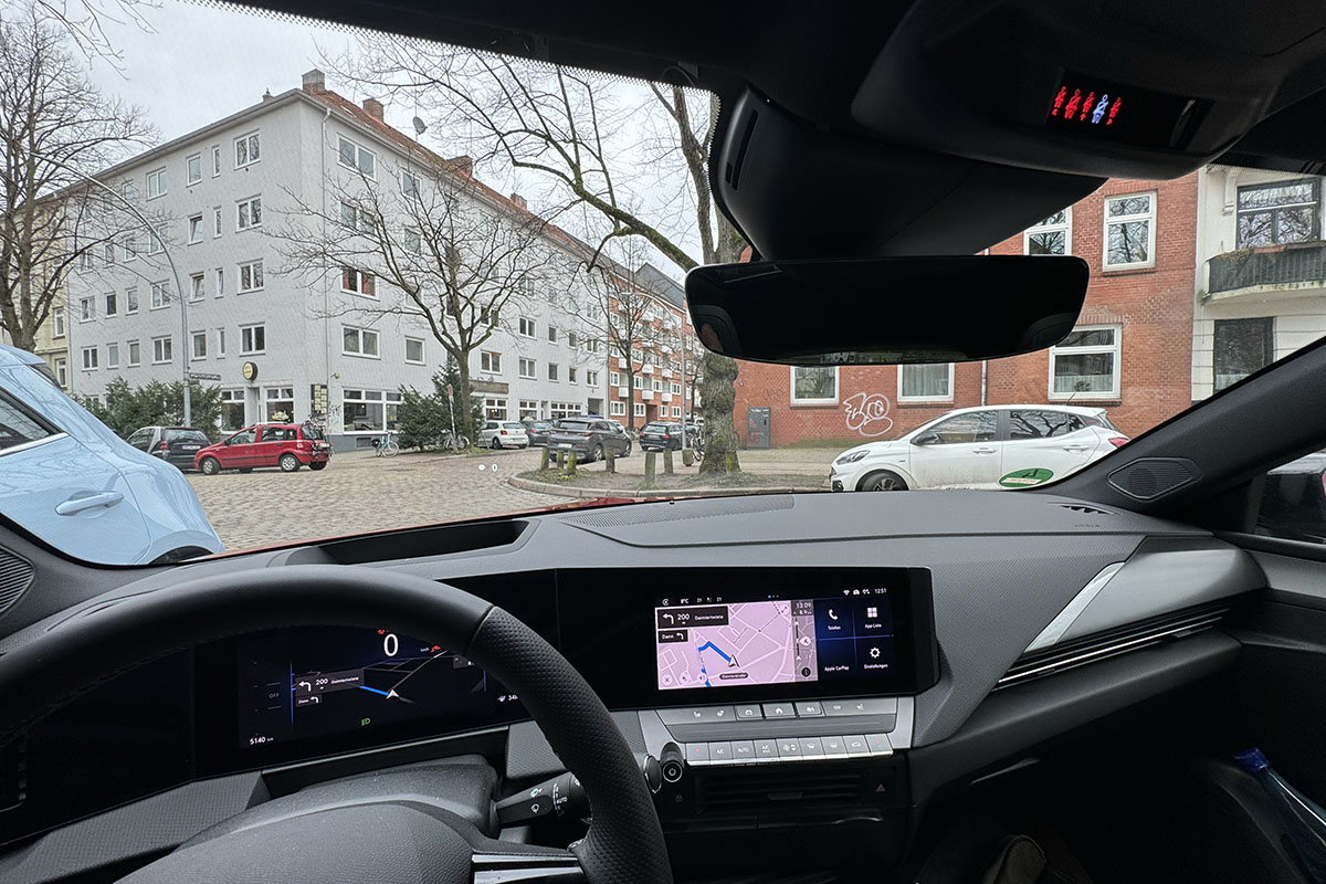 Blick durch die Frontscheibe des E-Autos Opel Astra Electric auf ein Wohnhaus in einer städtischen Umgebung.