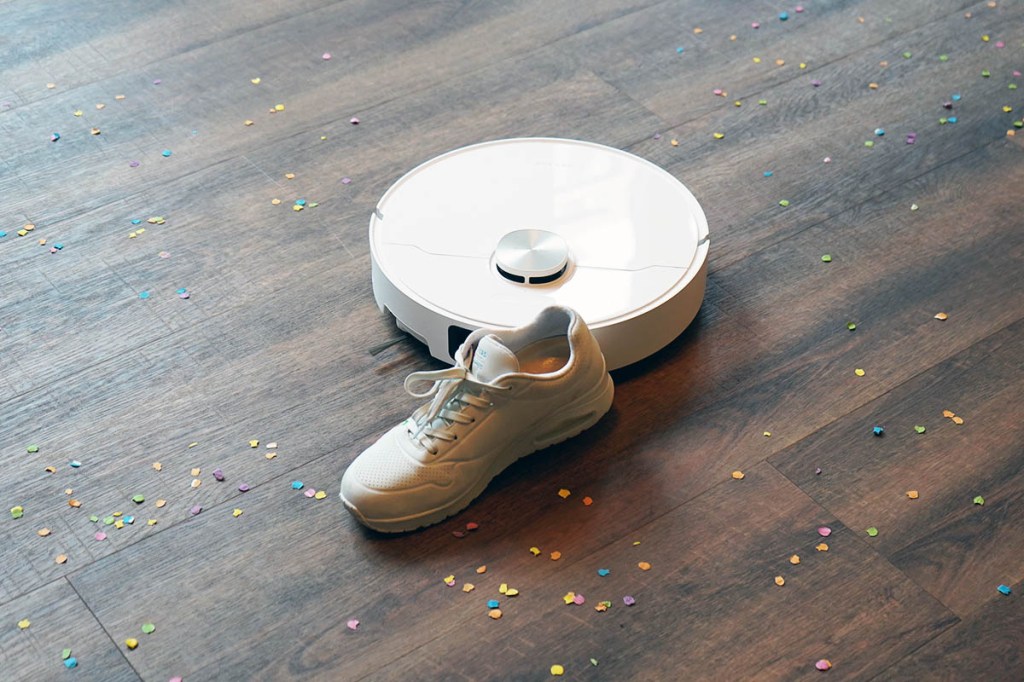 Der Dreame X40 Ulra Complete auf einem Holzboden mit Konfetti. Vor ihm liegt ein weißer Schuh.