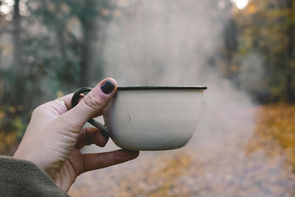 Frauenhand hält dampfende Kaffeetasse vor Waldstück in die Luft.