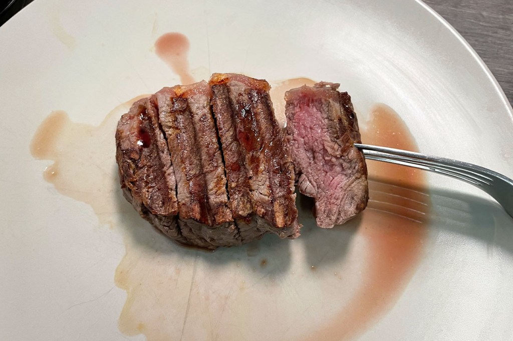 Ein Stück gegrilltes Steak auf einem hellen Teller. Es sind Grillmarkierungen zu sehen und eine Gabel öffnet das Fleisch an einer Schnittstelle.