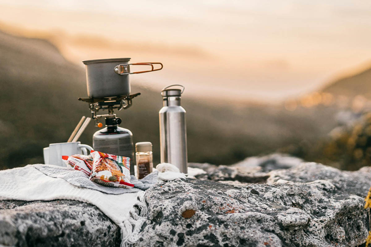 Ein Gaskocher und eine Trinkflasche stehen auf einem Stein vor der aufgehenden Sonne.