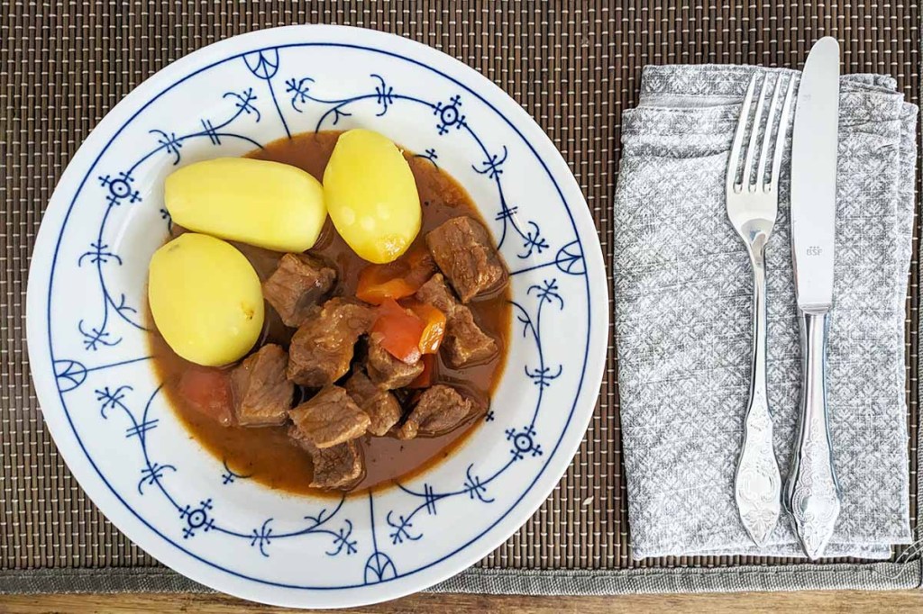 Gulasch mit Kartoffeln auf einem weiß-blauen Teller neben Besteck.