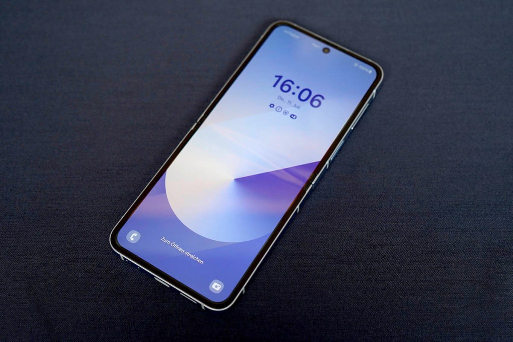Das faltbare Smartphone Samsung Galaxy Z Flip6 liegt mit eingeschaltetem Display auf einer dunkelblauen Fläche.
