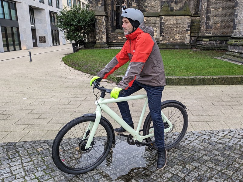 Mann mit grau-roter Jack Wolfskin Morobbia-Regenjacke auf Fahrrad, das in einer Pfütze auf einem gepflasterten städtischen Platz steht.