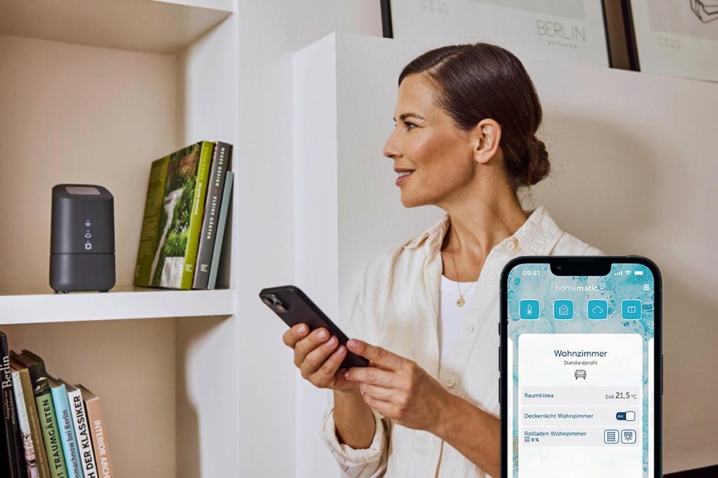Eine Frau steht mit ihrem Smartphone vor einem Regal, in dem die Home Control Unit steht.
