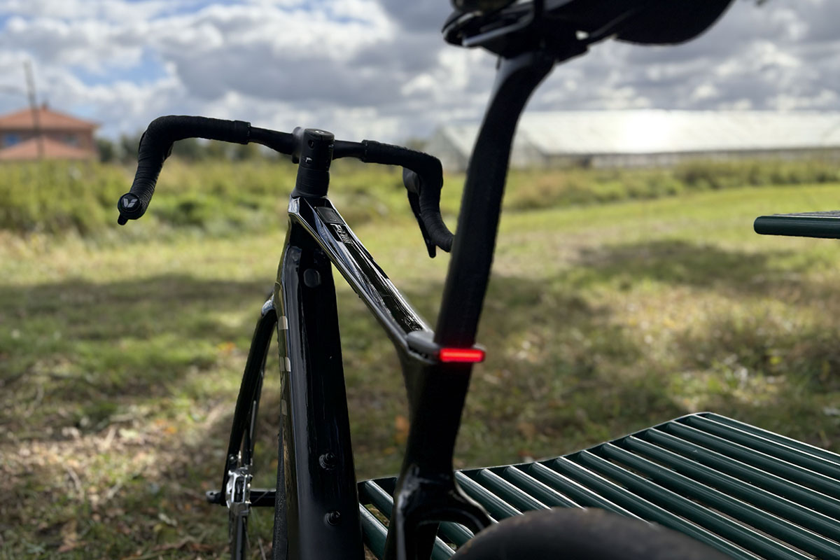 E-Rennrad von hinten fotografiert steht vor einem Feld, an der Sattelstütze ist ein eingeschaltetes Rücklicht zu sehen.