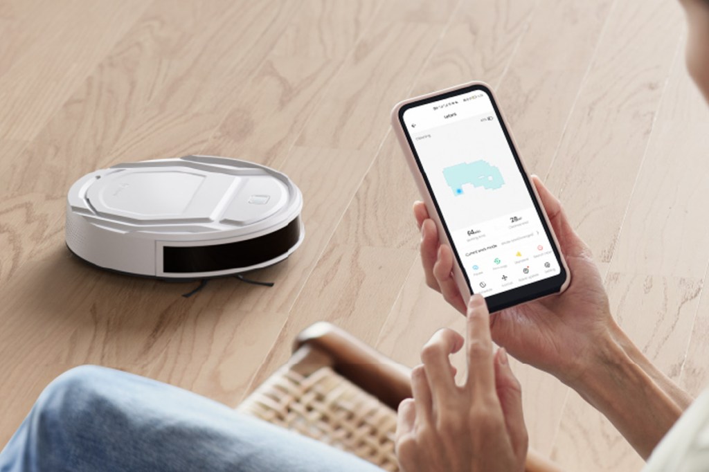 Eine Person guckt im Vordergrund des Bildes auf ein Smartphone. Dahinter fährt der Saugroboter auf Holzfußboden.