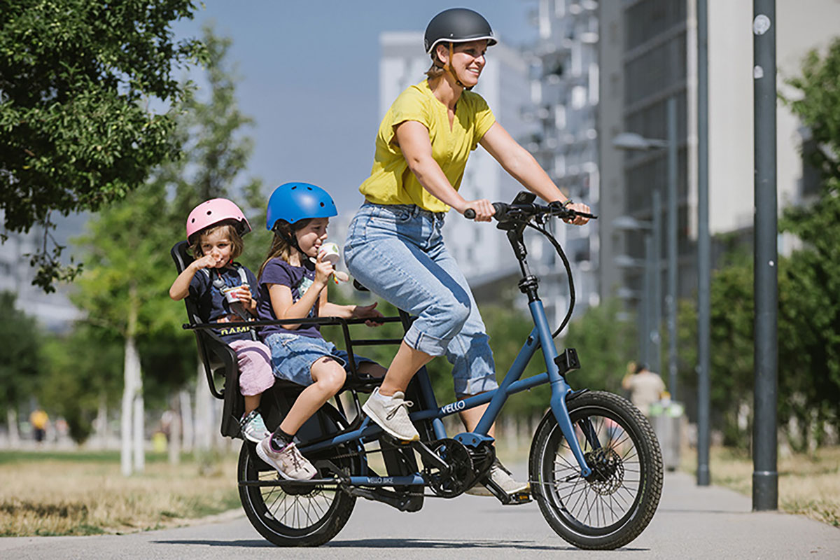 Frau fährtb auf klappbaren E-Cargo-Bike Vello Sub auf einer Straße ein einer Stadt. Auf dem Gepäckträger befinden sich zwei Kindersitze, in denen Kinder sitzen.