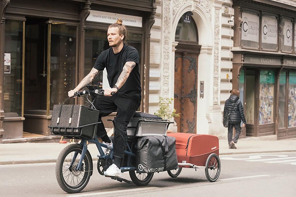 Mann fährt auf faltbaren Vello Sub-E-Cargo-Bike durch ein städtisaches Gebiet. Das kompakte Lastenrad ist dabei mit Gepäck auf einem Frontgepäckträger sowie einem Anhänger und Gepäck auf dem Heckgepäckträger reichlich mit Fracht beladen. 