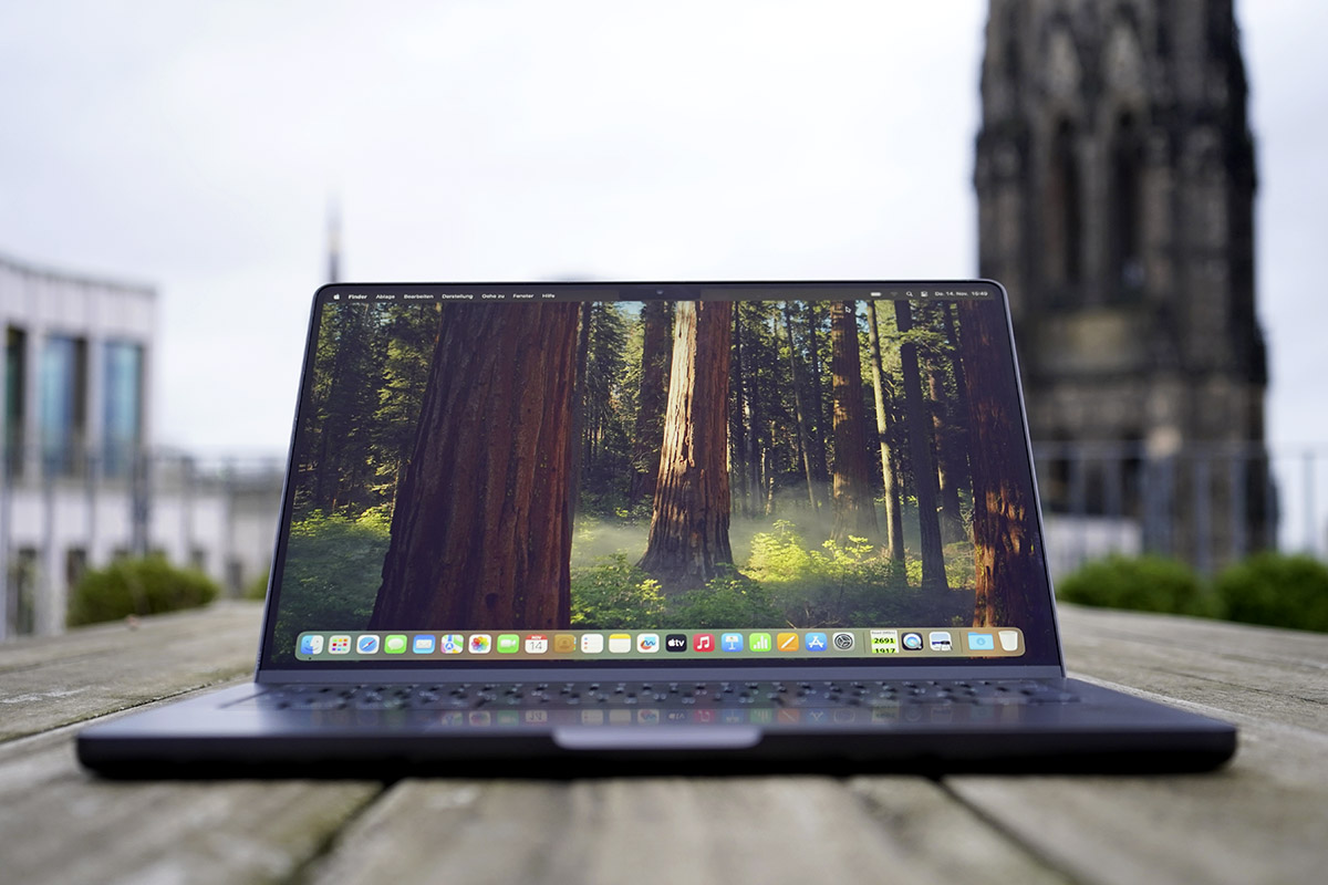 Ein Apple MacBook Steht aufgeklappt mit eingeschaltetem Bildschirm im Freien auf einem Holztisch.