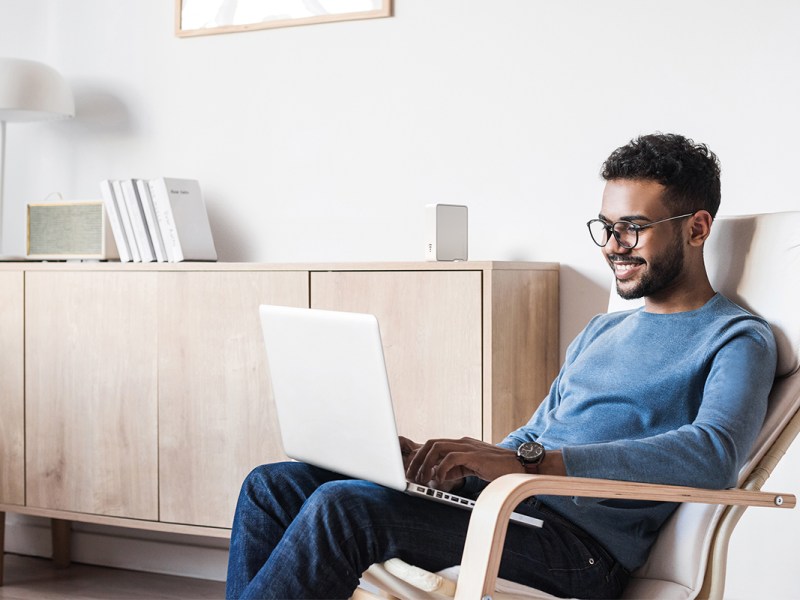 Ein Mann sitzt mit seinem Laptop in einem Sessel. Der Devolo Repeater steht im Hintergrund auf einem Sideboard.