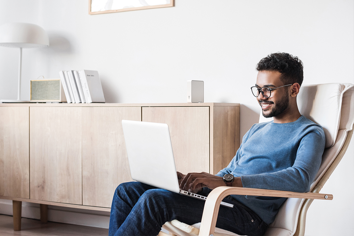 Ein Mann sitzt mit seinem Laptop in einem Sessel. Der Devolo Repeater steht im Hintergrund auf einem Sideboard.