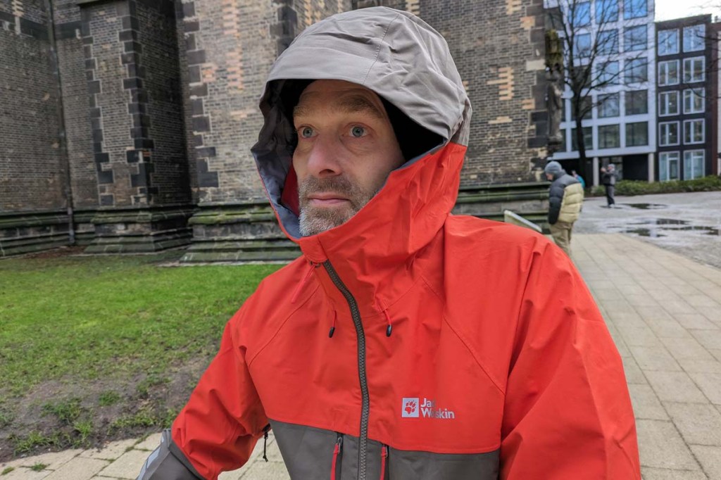 Person nah von vorne, trägt eine rot-braune Regenjacke und hat Kapuze auf