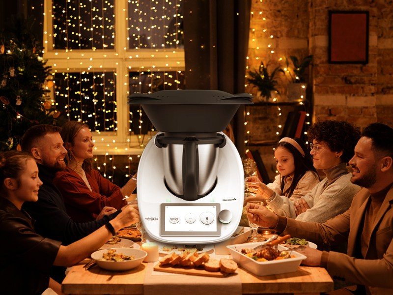 Eine Familie sitzt an einem Esstisch. Links ein Weihnachtsbaum, der Raum ist festlich geschmückt. Auf den Tisch ein Thermomix montiert.