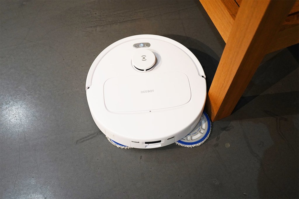 Der Ecovacs Deebot N30 Pro Omni von oben auf grauem Boden. Eine Wischplatte ist ausgefahren, um an einem Tischbein entlang zu wischen.