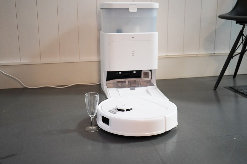 Ecovacs Deebot N30 Pro Omni fährt auf ein Glas zu. Dahinter ein Stuhl und die Station in wohnlicher Umgebung.