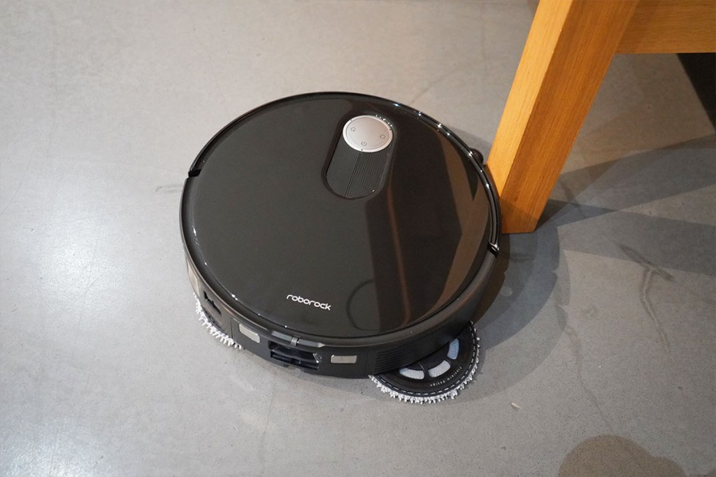 Der Roborock Qrevo Slim von oben auf grauem Boden. Eine Wischplatte ist ausgefahren, um an einem Tischbein entlang zu wischen.