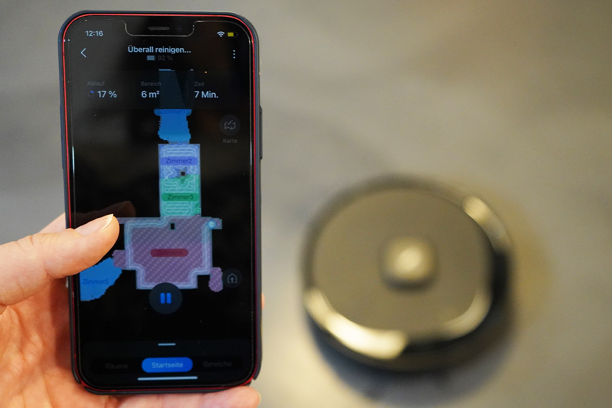 Eine Hand hält ein Smartphone in die Kamera. Darauf ist eine Saugroboter-App mit Karte zu sehen. Im Hintergrund fährt der Roboter.