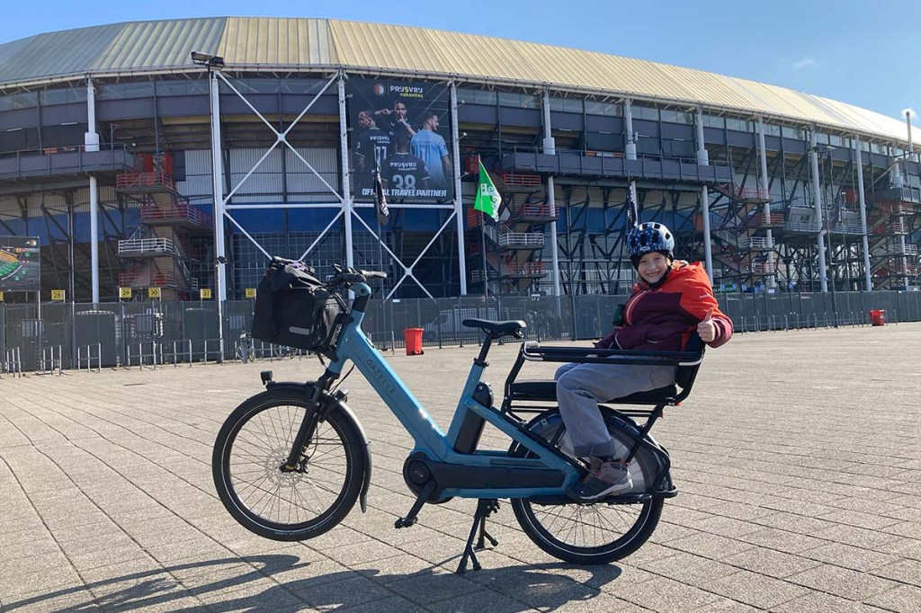 KInd auf dem Rücksitz eines E-Bikes sitzend, Fußballstadion im Hintergrund